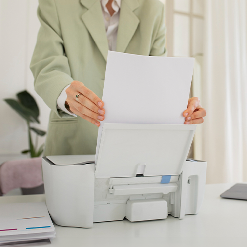 A Woman in a Suit Is Holding a Paper in Front of a Printer. She Is Using Check Print Software Free to Efficiently Manage and Print Checks.