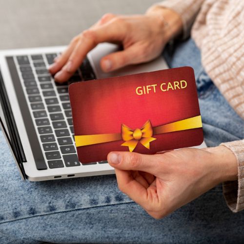 A Woman Holding a Gift Card in Front of Her Laptop. She Is Send to Gift Card Online to Anyone in Her Payee List