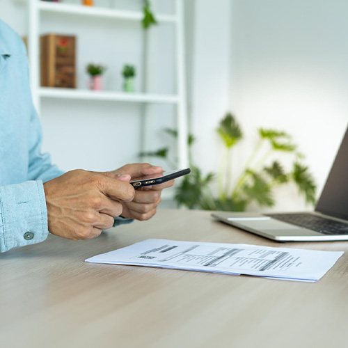 A Person Holding a Phone Seated at a Desk Engaged in Bill Payment Over a Document Next to a Laptop.