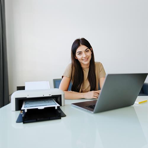 Smiling Woman Using Check Writing Software on Her Laptop at a Tidy Office Desk with a Printer Beside Her.