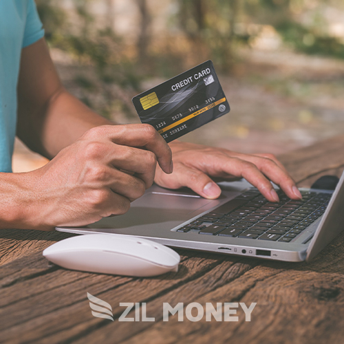 Man Sitting at a Wooden Table Outdoors Using a Laptop While Holding a Credit Card in One Hand. He Is Engaged in a Task, Looking Through Options to Rent Pay by Credit Card