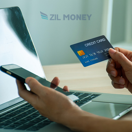 A Person Holding a Credit Card in One Hand and a Smartphone Pay Bills with Credit Cards, While Sitting in Front of an Open Laptop. the Logo "Zil Money" Appears at the Top of the Image.