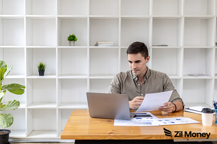 A Man Reviews Documents While Working On A Laptop At A Clean Desk, Highlighting Organized And Efficient Invoice Management.